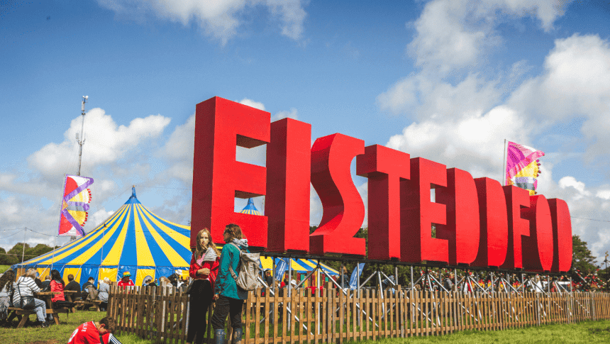 Photo showing the eisteddfod site on a bright day with clear blue skies. The huge eisteddfod letters are in he foreground with the main pavillion in the background.