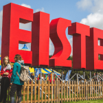 Photo showing the eisteddfod site on a bright day with clear blue skies. The huge eisteddfod letters are in he foreground with the main pavillion in the background.