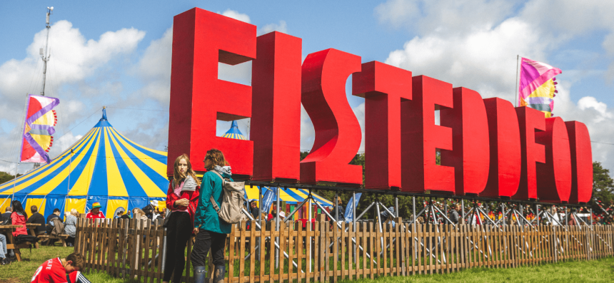 Photo showing the eisteddfod site on a bright day with clear blue skies. The huge eisteddfod letters are in he foreground with the main pavillion in the background.