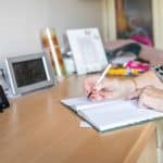 Older person leaning on a counter writing in a notebook, with a smart meter display on the left