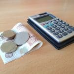 A small pile of silver coins on top of a folded £10 note next to a calculator on a desk
