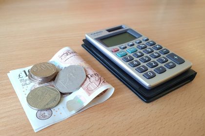A small pile of silver coins on top of a folded £10 note next to a calculator on a desk