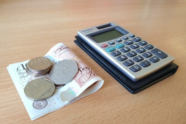 A small pile of silver coins on top of a folded £10 note next to a calculator on a desk