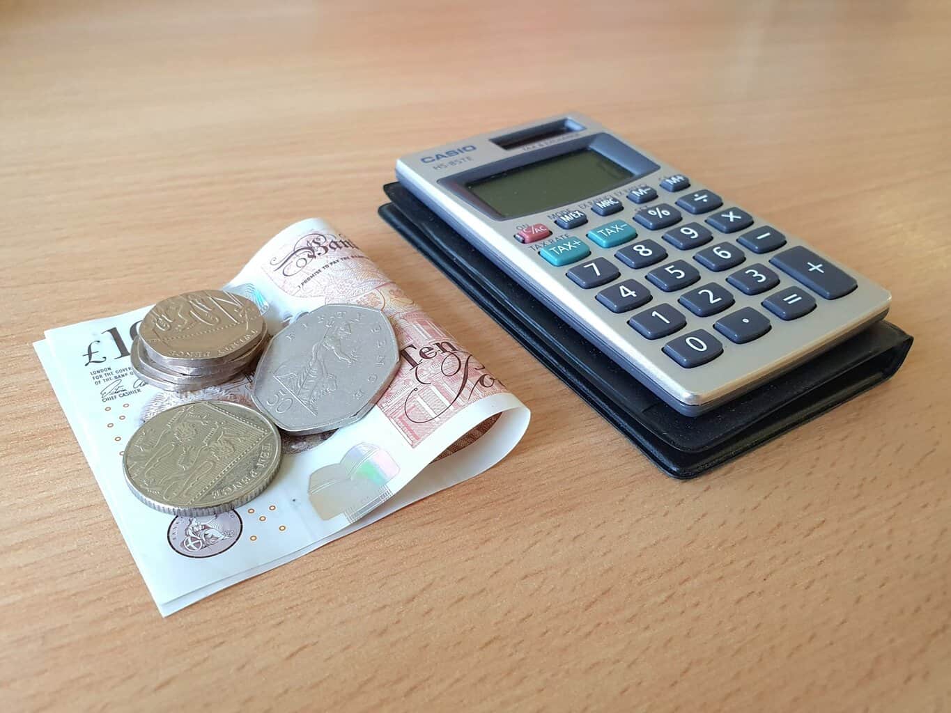 A small pile of silver coins on top of a folded £10 note next to a calculator on a desk